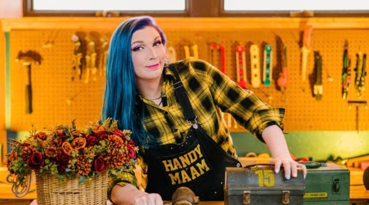 Mercury Stardust leans over a wooden table with a toolkit, holding a hammer, and wearing an apron that say "HANDY MA'AM."