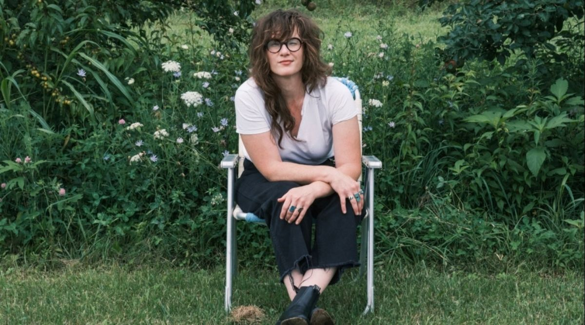 S.G. Goodman sits in a folding lawn chair in a field of wildflowers, wearing a white T-shirt, black wide-leg jeans and black boots.