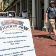 A Whiskey Row Walking Tour sandwich board sign on a brick sidewalk in downtown Louisville as a man in a sun hat walks past.