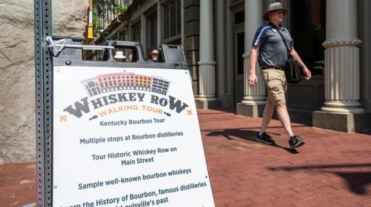 A Whiskey Row Walking Tour sandwich board sign on a brick sidewalk in downtown Louisville as a man in a sun hat walks past.