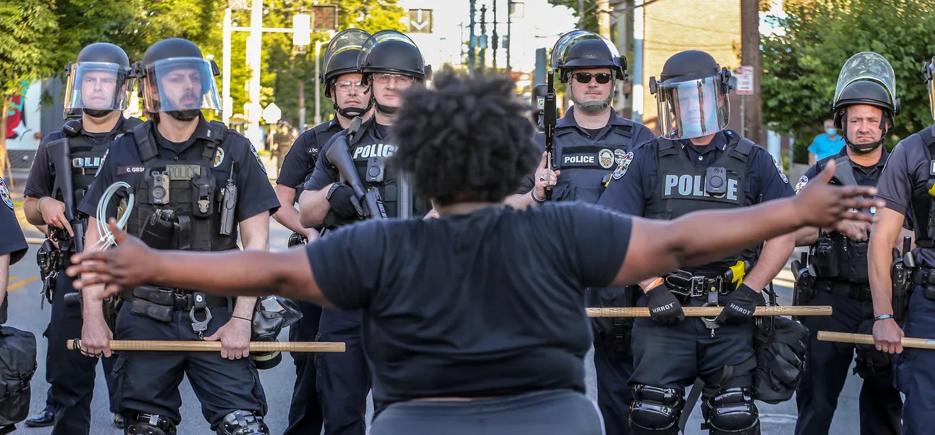 A protester stands with arms outstretched facing a line of police officers in riot gear holding batons.