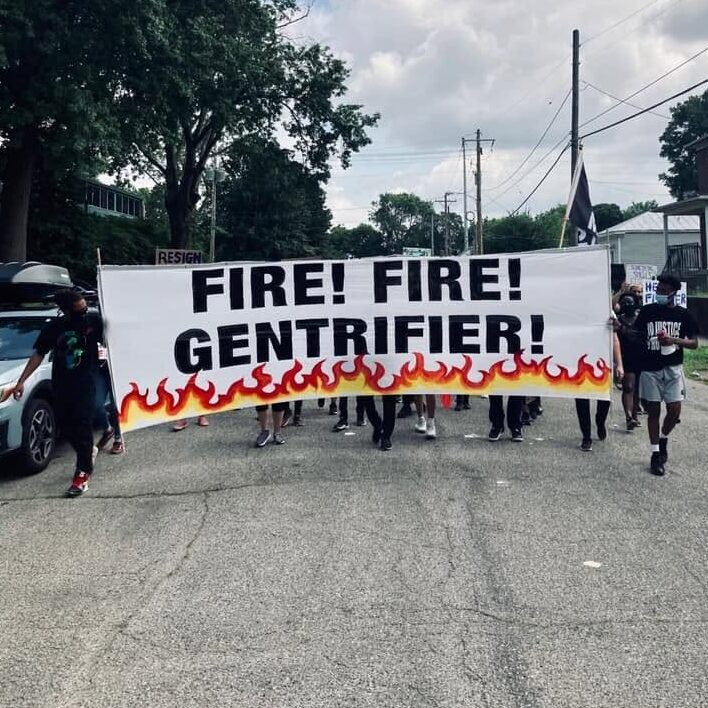 Marchers carry a large banner reading "Fire! Fire! Gentrifier!" down a street in summer 2020.