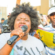 A speaker holds a microphone and addresses a crowd at an outdoor rally as people gather behind her.