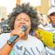 A speaker holds a microphone and addresses a crowd at an outdoor rally as people gather behind her.