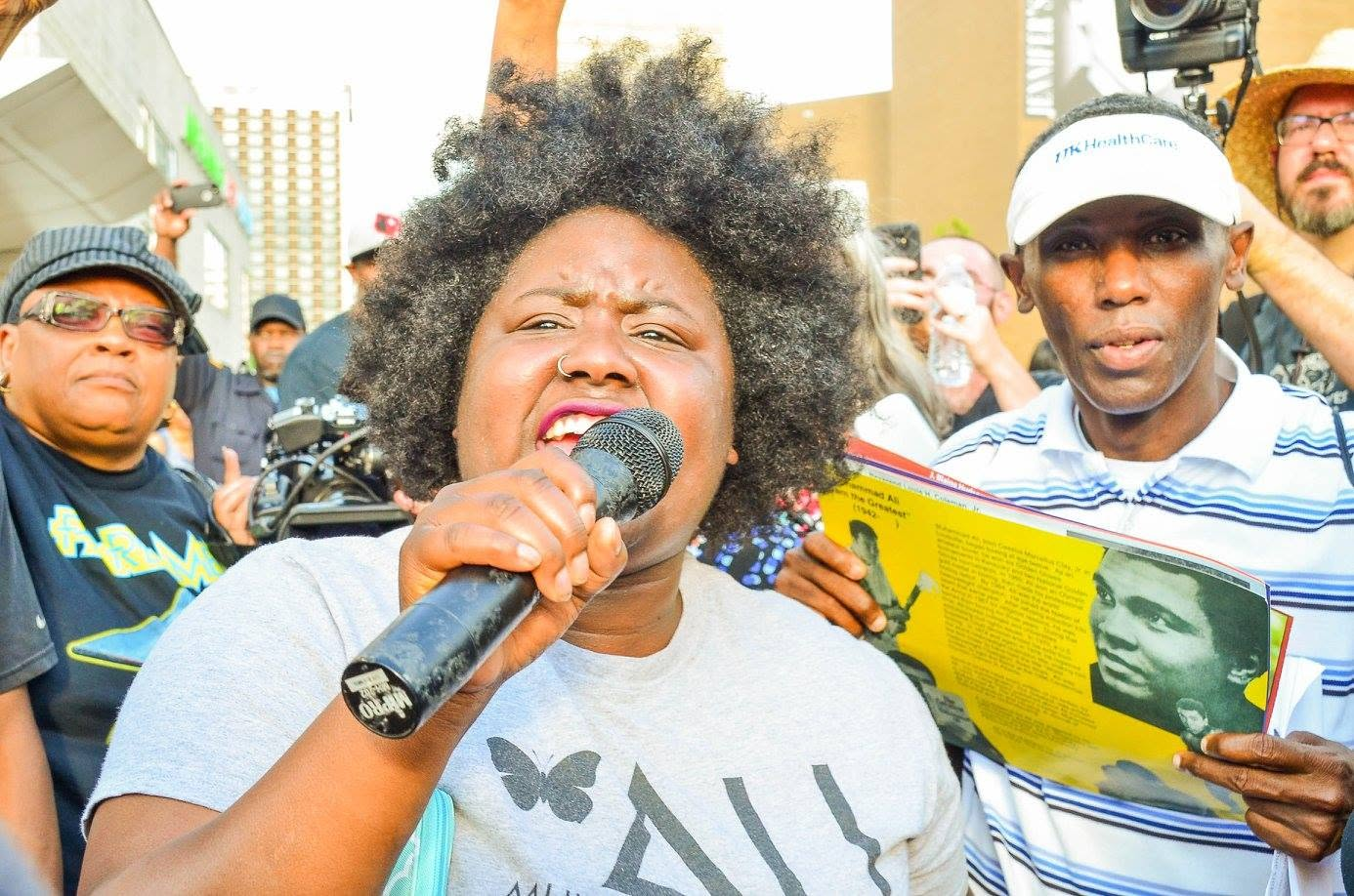 A speaker holds a microphone and addresses a crowd at an outdoor rally as people gather behind her.