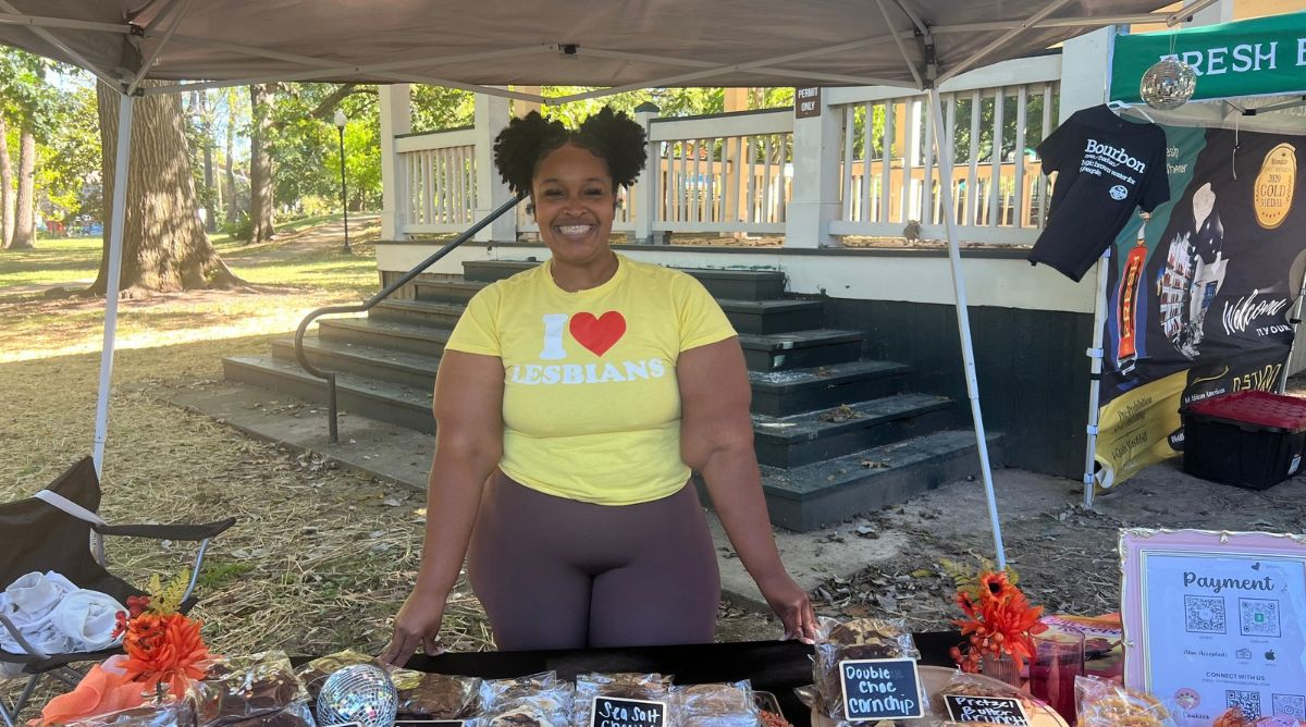 A landscape image of a woman with space buns standing behind a table, under a tent, vending for an event. She is wearing a yellow t-shirt that says "I [heart] Lesbians"