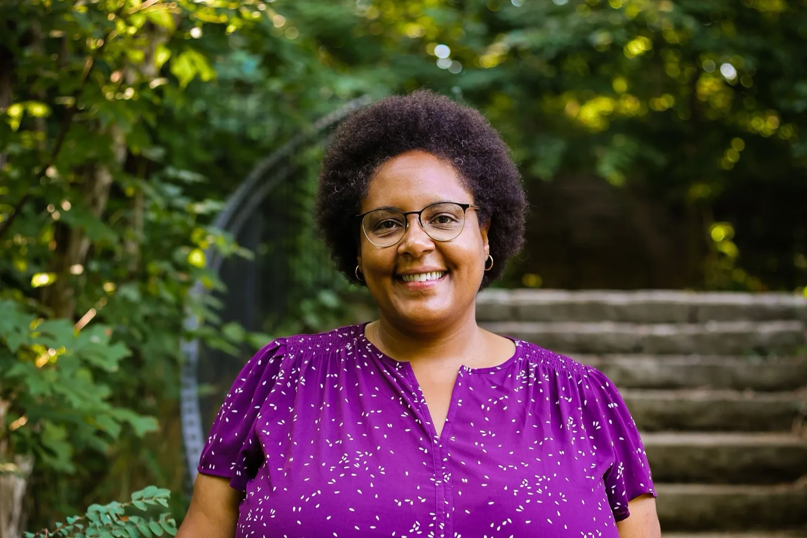 Alicia Hurle, co-executive director of Common Ground Kentucky, smiling outdoors in a purple top in front of stone steps and trees