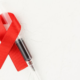 A red AIDS ribbon lays on a white background with a syringe of blood next to it.
