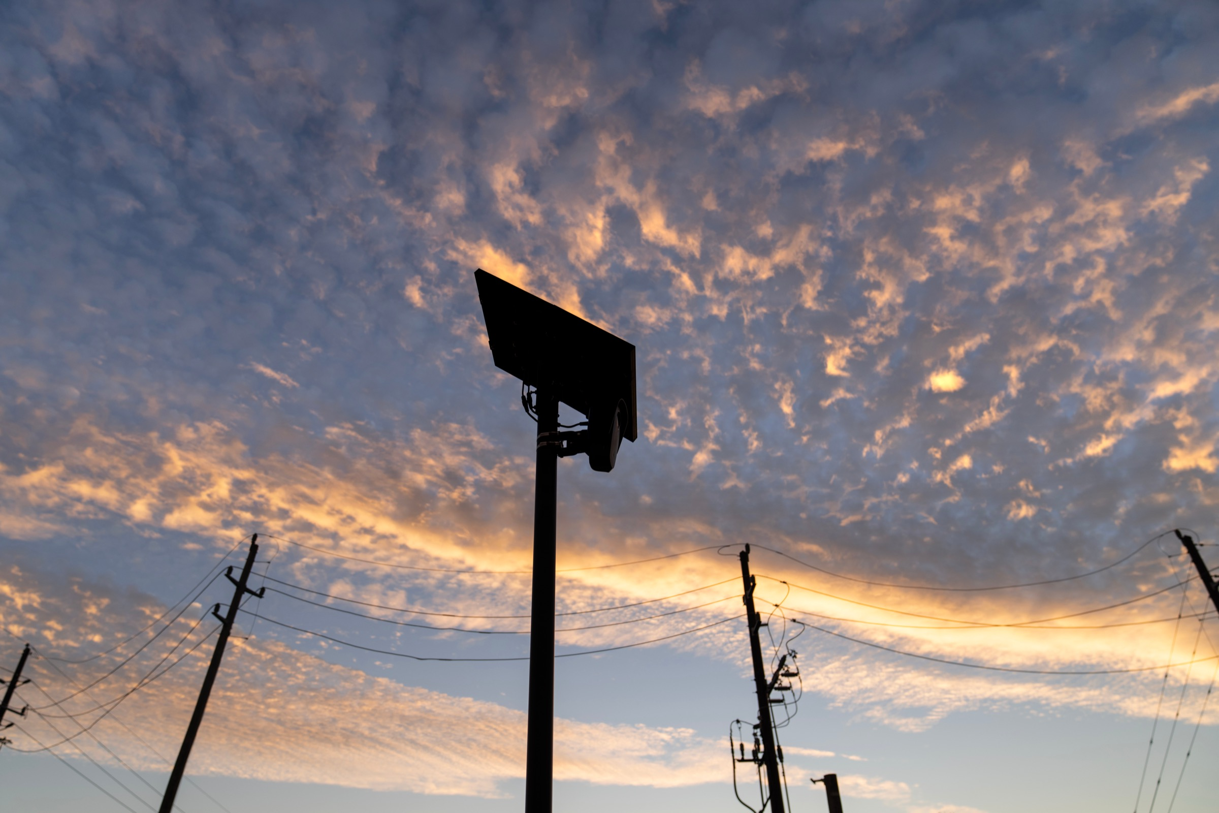 A Flock license plate reader camera on a pole against a cloudy sunset sky, with power lines in the background.