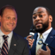 Andy Barr in a suit and striped tie smiles at a podium, beside Charles Booker speaking into a microphone, on a dark background.