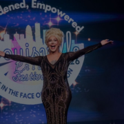 A drag performer in a sequined black gown raises their arms on stage in front of the 2026 Louisville Pride Festival sign reading "Emboldened, Empowered."