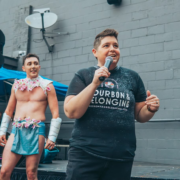 Missy Spears speaks into a microphone at an outdoor event, with drag performers standing behind her.