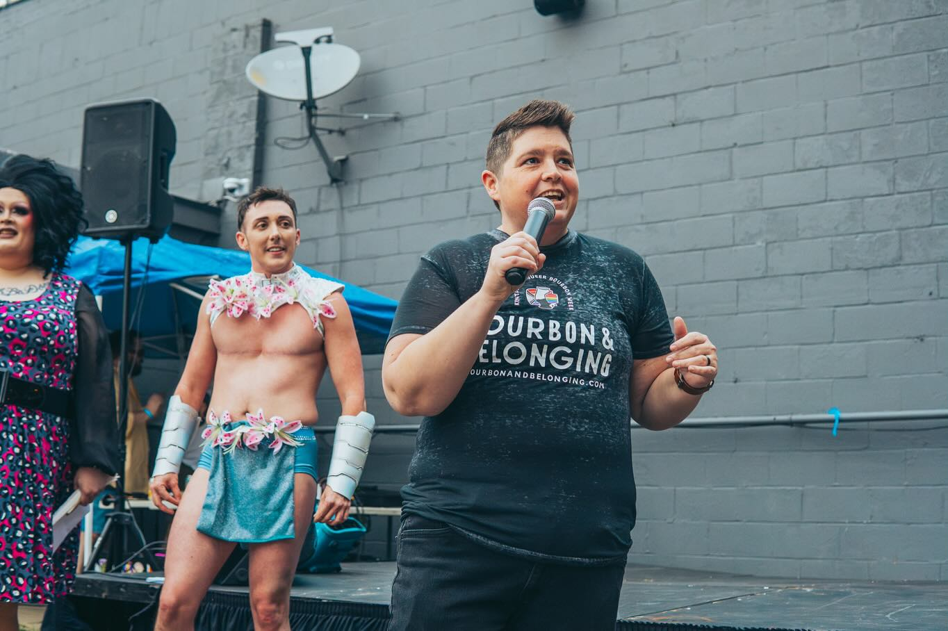 Missy Spears speaks into a microphone at an outdoor event, with drag performers standing behind her.