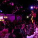 A packed Louisville crowd applauds two Time Slip Theatre performers standing on a runway stage beneath a cluster of pink and purple disco balls.