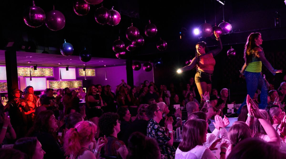 A packed Louisville crowd applauds two Time Slip Theatre performers standing on a runway stage beneath a cluster of pink and purple disco balls.