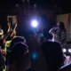 A performer in a striped shirt sings on an outdoor stage at night while the crowd raises their hands.